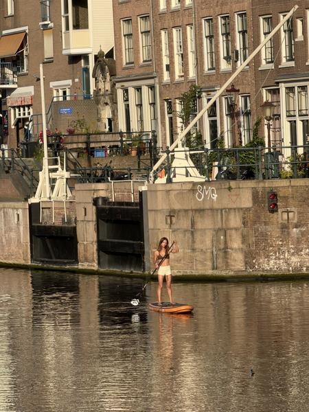 A person is stand-up paddleboarding on the tranquil canal in Oudeschans, Uilenburg, Amsterdam, bathed in warm afternoon sunlight